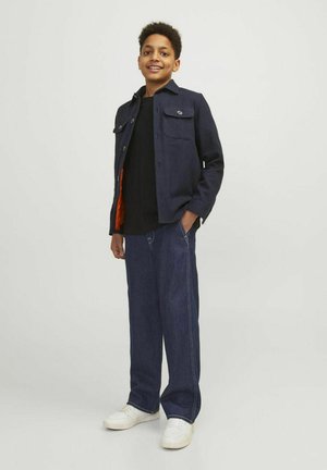Veste en denim bleu marine avec fermeture à boutons, deux poches sur la poitrine et doublure orange, associée à un t-shirt noir et un pantalon en denim fluide. Baskets blanches.