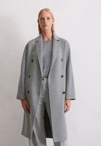Hellgrauer Wollmantel mit doppelt geführter Knopfleiste und großen schwarzen Knöpfen, ausgestattet mit einem Reverskragen und einer lockeren Passform, kombiniert mit einem grauen Strickpullover und -hosen.