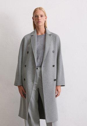 Manteau en laine gris clair à double boutonnage avec de grands boutons noirs, dotée d'un col cranté et d'une coupe décontractée, associée à un pull en maille gris et à un pantalon.