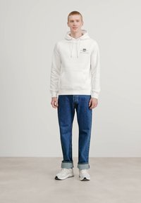 Haut à capuche blanc avec une poche avant et des cordons, associé à un jean bleu et des baskets blanches, se tenant sur un fond neutre.