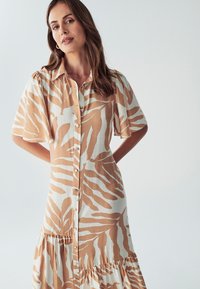 Košilové šaty v slonovinovej farbe s svetlohnedým vzorom listov; majú krátke rukávy, volánovú sukňu a voľný strih.