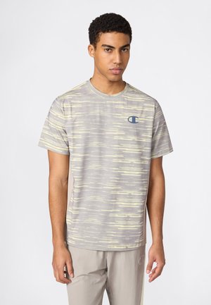 Jonge man die een grijs-gele gestreepte korte mouw t-shirt en lichtgrijze broek draagt, staand tegen een eenvoudige witte achtergrond.