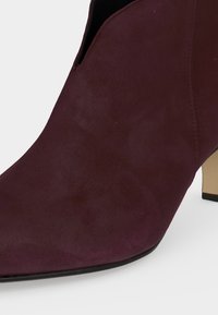Gros plan sur une bottine en daim bordeaux avec un talon bloc beige et une semelle extérieure noire sur fond clair.