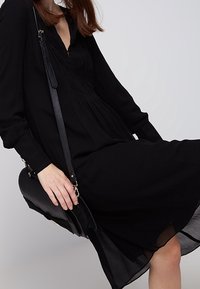 Czarna sukienka z długimi rękawami, dekoltem w kształcie litery V i marszczonymi detalami. Zestawiona z czarną torebką na ramię. Gładki materiał, długość do połowy łydki, luźny krój.