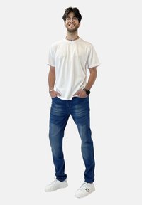 Camiseta de algodón blanca, jeans azul oscuro con ligero desgaste y zapatillas blancas con detalles en negro. El modelo está de pie con las manos en los bolsillos.