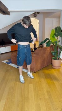 Camisa preta de mangas longas, calções de brim com vários bolsos, ténis cinzentos e um chão de madeira num ambiente de sala de estar.