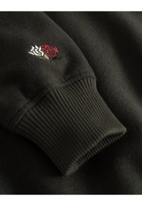Felpa nera con polsini a coste, decorata con design di rose rosse e bianche ricamate. La texture morbida e la vestibilità strutturata sono evidenti.