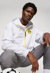 Homme en veste blanche de Borussia Dortmund et pantalon gris s'accroupissant à côté d'un ballon de football sur un fond neutre.