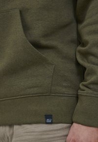 Olivengrüner Sweatshirt mit einer Vordertasche, gerippten Bündchen und Saum. Verfügt über ein kleines schwarzes Etikett auf der unteren linken Seite. Weicher Stoff.