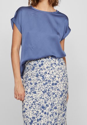 Vrouw draagt een blauwe korte mouwen blouse ingestopt in een witte rok met blauw bloemmotief, staand tegen een effen achtergrond.