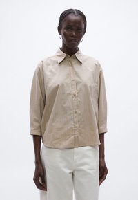 Cămașă bej cu dungi, cu nasturi, guler larg, mâneci trei sferturi și nasturi în față, purtată cu pantaloni albi. Textură netedă a țesăturii.