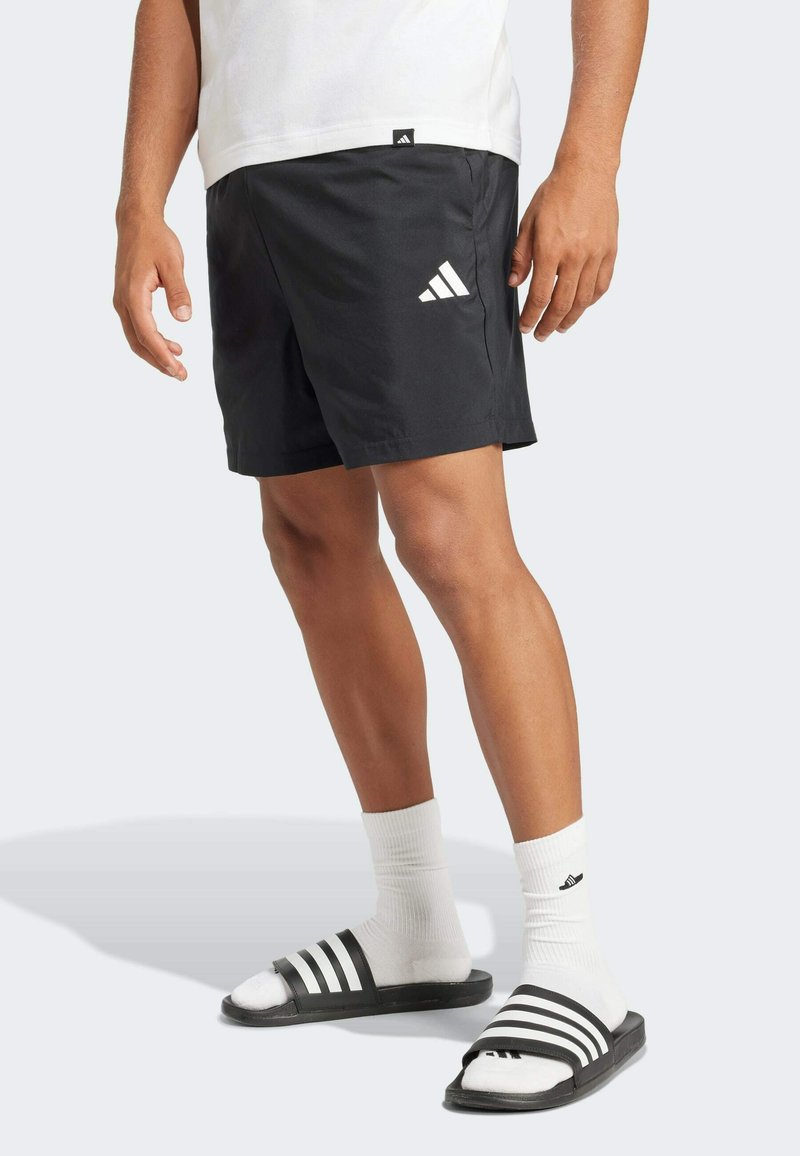 Mustat urheiluShortsit, joissa on joustava vyötärö, valkoisella Adidas-logolla, yhdistettynä valkoisiin sukkiin ja mustiin liukuessandaaleihin, joissa on valkoiset raidat.