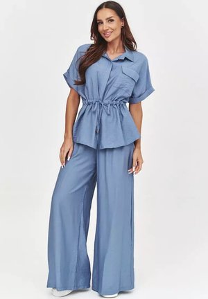 Uśmiechnięta kobieta ubrana w luźny, jasnoniebieski top z krótkim rękawem ze ściągaczem w talii oraz pasujące szerokie spodnie na jednolitym tle.