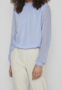 Světle modrá halenka s dlouhým rukávem a strukturovaným zigzagovým vzorem, nabíraným výstřihem a elastickými manžetami, doplněná o krémové kalhoty.