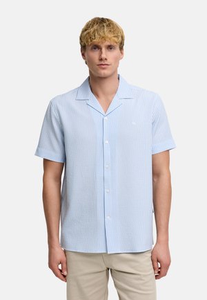 Jeune homme aux cheveux blonds courts portant une chemise à manches courtes rayée verticalement bleu clair et blanc et un pantalon beige.