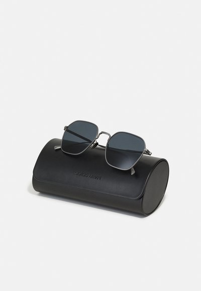 Ochelari de soare din metal cu lentile întunecate, formă pătrată, așezați pe un toc rotund din piele neagră, embosat cu "Giorgio Armani."
