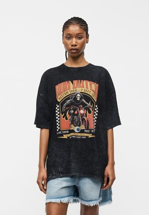 Νεαρός με πλεξούδες, που φοράει μαύρο, φθαρμένο, φαρδύ μπλουζάκι με ένα γκραφικό κρανίου μοτοσικλετιστή και κοντά τζιν σορτς.