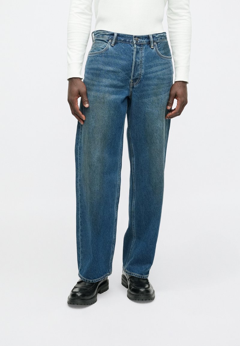 Oseba v modrih ozkih hlačah, črnih vezalkah in dolgo rokavno beli majici, ki stoji pred enobarvno svetlo ozadje.