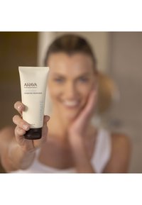 Mascarilla hidratante AHAVA en un tubo blanco con texto azul, con una base negra. El producto es sostenido por una persona frente a un fondo desenfocado.