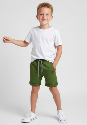 Uśmiechnięty chłopiec z kręconymi włosami, ubrany w białą koszulkę, zielone szorty ze sznurkiem oraz białe trampki, stojący z jedną ręką w kieszeni.
