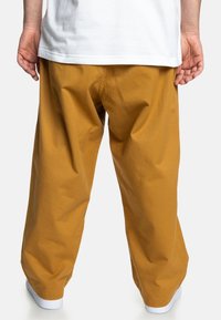 Volné kalhoty v barvě hořčicové žluté z bavlněného materiálu; mají jednoduchý design s minimem viditelných švů a bílé obutí.