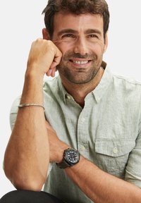 Zwart digitaal horloge met groene rubberen band, grote wijzerplaat die cijfers en meerdere wijzerplaten weergeeft. Draag het met een lichtgroen overhemd met knopen.