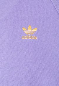 Purple fabric with a smooth texture features a yellow embroidered Adidas logo and trefoil design at the lower corner, indicating brand identity.