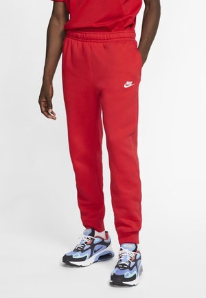 Mężczyzna w jasnoczerwonych spodniach dresowych Nike i wielokolorowych butach Nike Air Max, stojący na tle jednolitego białego tła.