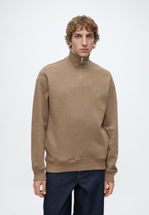 Junger Mann trägt einen beigen Pullover mit Viertel-Reißverschluss und dunkelblaue Jeans, steht vor einem schlichten hellen Hintergrund.