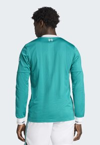 Maillot de sport à manches longues en teal avec un tissu texturé, col blanc et poignets rayés. Il présente un logo au dos et des accents contrastés sur les manches.