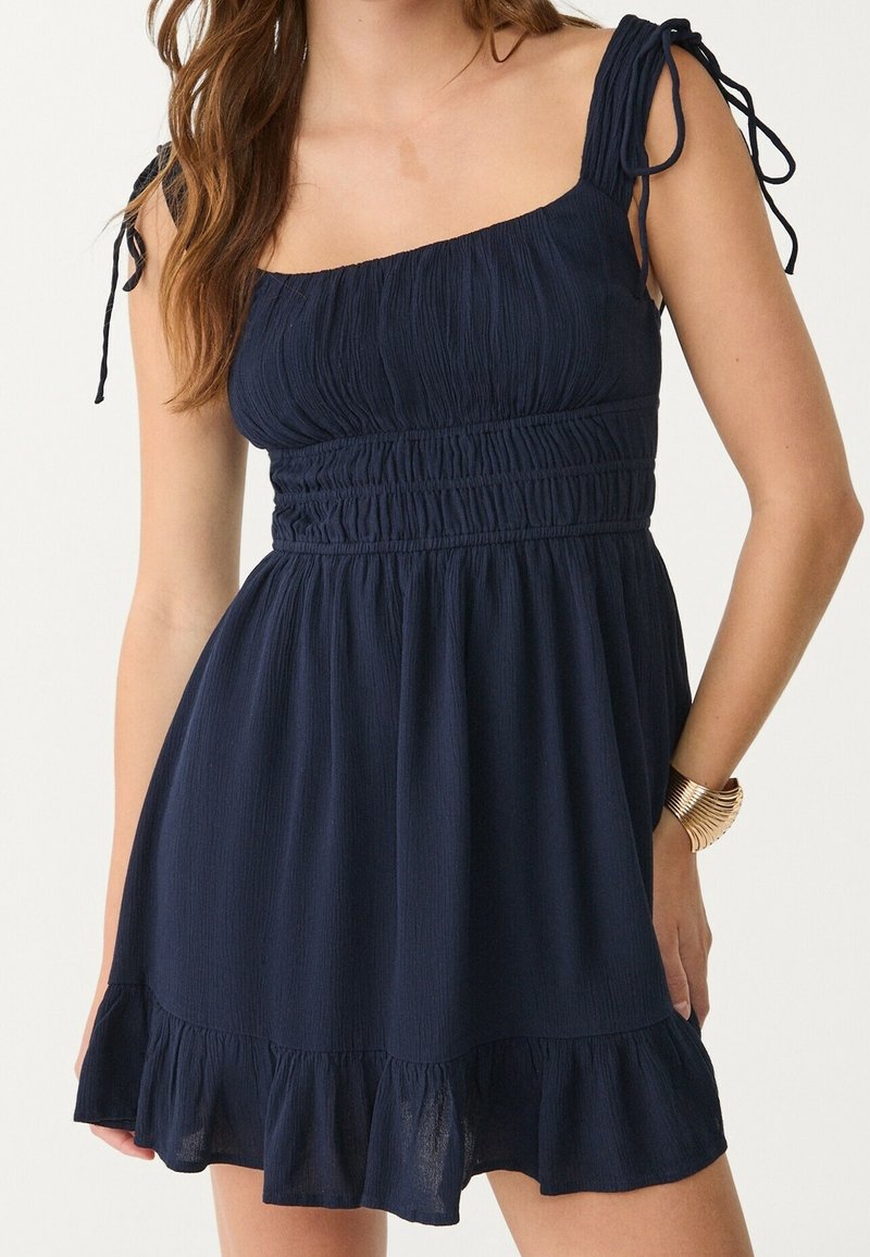 Femme portant une robe sans manches bleu marine avec un corsage froncé, des bretelles à nouer sur les épaules et un ourlet à volants, accessoirisée d'un bracelet en or.