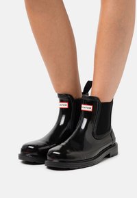 Zwarte rubberen enkelboots met elastische zijkanten, ronde neus en een stevige zool. Bevat een rood-wit logo op de bovenste sectie.