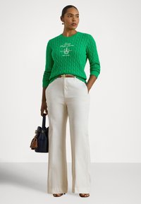 Zielony sweter w serek z dzianiny o grubym splocie, z okrągłym dekoltem, noszony na beżowych szerokich spodniach. Uzupełniony granatową torbą z złotymi dodatkami.