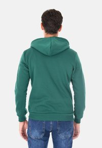 Felpa verde realizzata in morbido tessuto, con un grande cappuccio, polsini e orlo a costine. Indossata sopra dei jeans in denim, senza loghi o fantasie visibili.