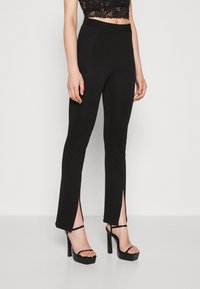 Pantalons noirs ajustés à taille haute, avec une fente à l'ourlet aux chevilles. Le matériau a une texture lisse et un design élégant.