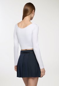 Biała krótka bluzka z długim rękawem zestawiona z granatową spódnicą plisowaną z cienkimi pionowymi paseczkami i czarnym pasem, widziana z tyłu.