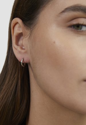 Nahaufnahme des linken Ohrs einer Person mit kleinem silbernem Creolenohrring und langen braunen Haaren vor einem schlichten Hintergrund.