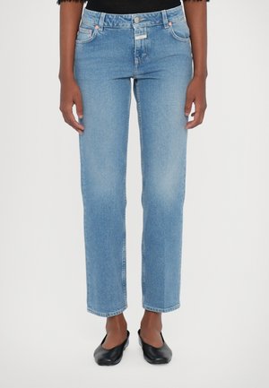 Personne portant un jean droit bleu clair et des chaussures noires sans lacets, debout devant un fond blanc uni.