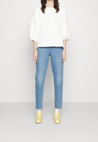 Femme portant un haut à manches bouffantes blanches, un jean bleu clair, des chaussettes blanches et des mocassins jaunes, se tenant contre un fond clair.