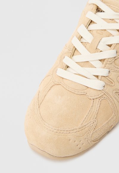 Sneaker in suede beige con tomaia texturizzata, cuciture a vista e lacci color crema. Presenta una punta arrotondata e dettagli laterali forati.