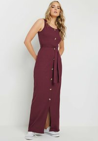 Robe maxi bordeaux avec texture côtelée, boutons sur le devant, taille ceinturée et fente sur le côté, associée à des baskets blanches.