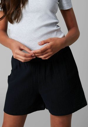 Femme portant un t-shirt à manches courtes côtelé gris clair et un short noir texturé et ample, sur un fond uni.