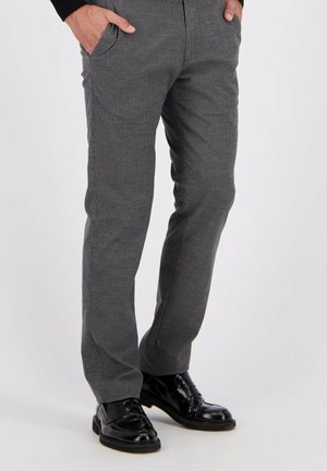 Man in slim-fit grijze pantalon en glanzende zwarte leren schoenen, staand met handen in zijn zakken op een effen achtergrond.