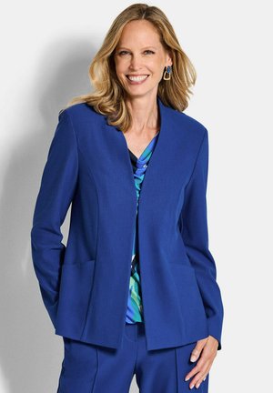 Lachende vrouw met blond haar, gekleed in een blauw pak en een patroonrijke blauwgroene top, staand tegen een lichtgrijze achtergrond.