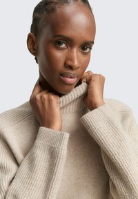 Beżowy sweter dzianinowy z wysokim, prążkowanym kołnierzem i szerokimi rękawami. Tekstura wydaje się miękka i przytulna, z delikatnym wzorem.