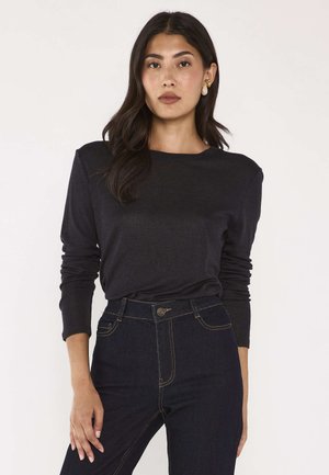 Donna con lunghi capelli scuri che indossa un top nero a maniche lunghe e jeans scuri a vita alta, in piedi di fronte a uno sfondo semplice.