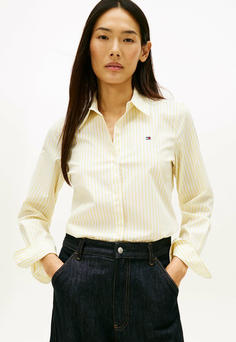 Lichtgele overhemd met witte verticale strepen en een klein logo op de borst; gecombineerd met donkere denim broek.