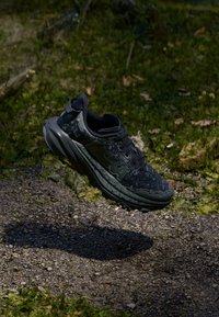 Black athletic shoe with textured upper, featuring a logo on the side, cushioned sole, and a sturdy lace-up design, elevated above ground.