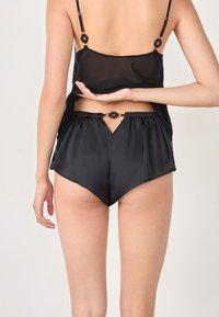 Modèle portant un haut noir transparent et un short satin assorti, vu de dos, mettant en avant un détail de découpe unique à la taille.