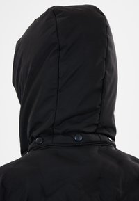 Svart pufferjacka med huva, som har en slät yta, sydda sömmar och tryckknappar vid halslinningen. Enkel design och funktionell form.